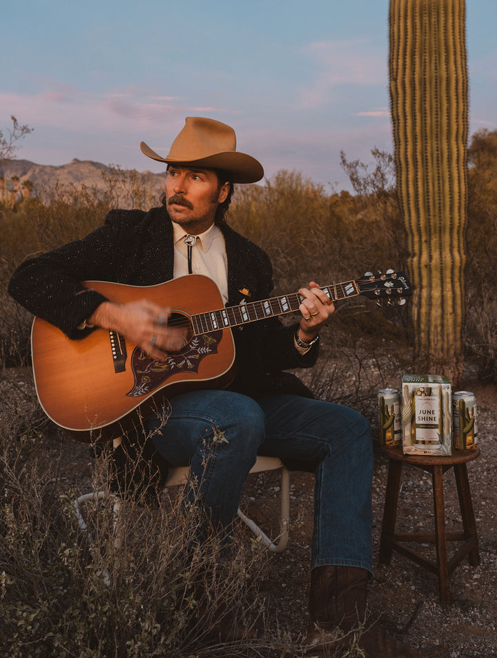 Mark Wystrach sitting in the desert among Saguaro cacti wiht his guitar, a cowboy hat, and a carton of Desert Cooler