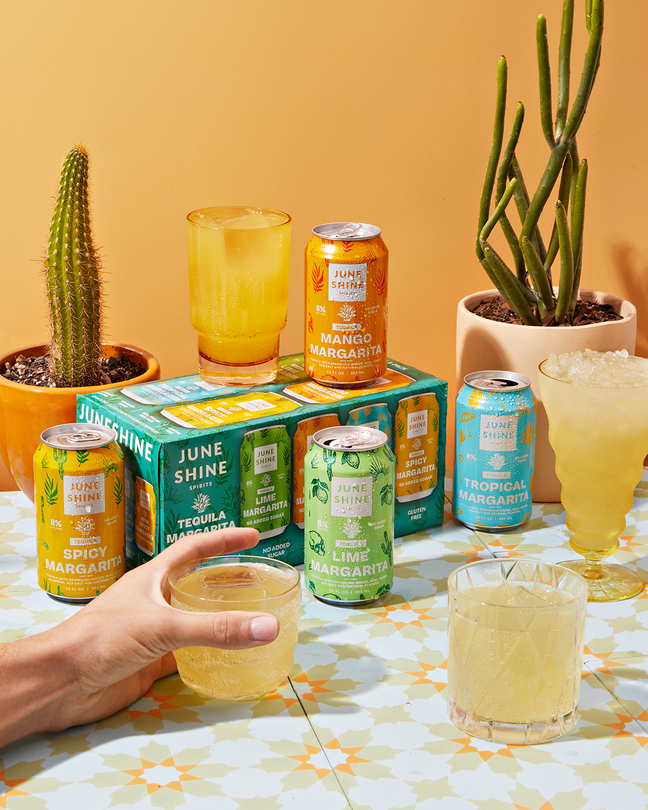 Margarita 8pk with 4 cans: Spicy Margarita, Lime Margarita, Mango Margarita, and Tropical Margarita all in cans and glasses in a Mexican setting