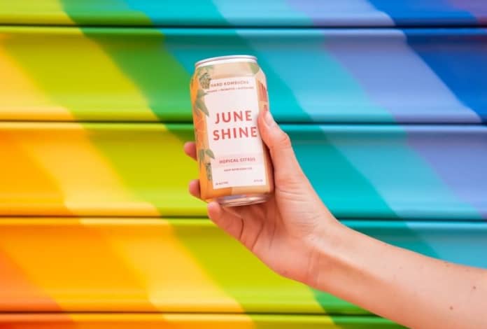A hand holding a JuneShine can against a rainbow background