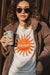 Photograph of woman wearing cream colored shirt with dark orange sun that reads "Shine" and also wearing corduroy jacket, sunglasses and holding can of june shine.