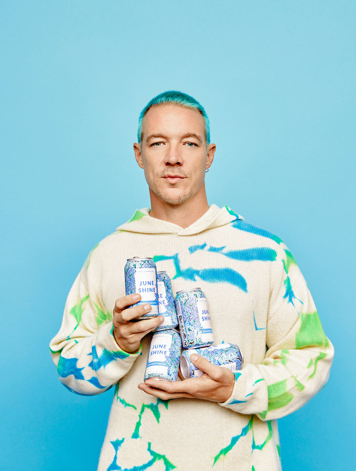 Diplo with blue hair and tie dye sweatshirt holding 5 unopened cans of Permanent Vacation