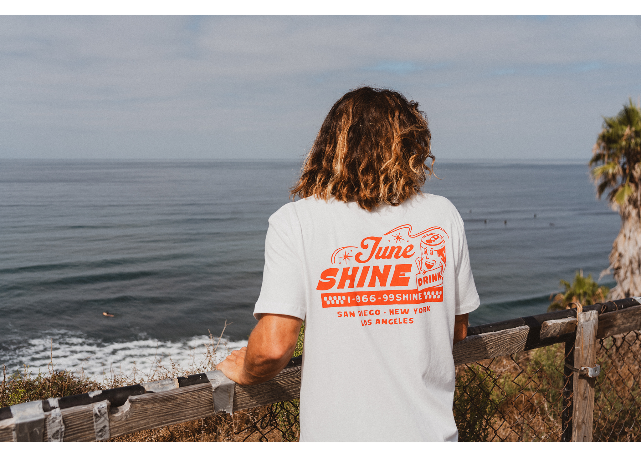 Long haired man at the beach wearing White tshirt with red writing that reads: JuneShine, 1-866-99SHINE San Diego New York Los Angeles with a popped can