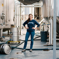 Smiling brewer standing in brewing facility with hoses, kegs, and barrels.