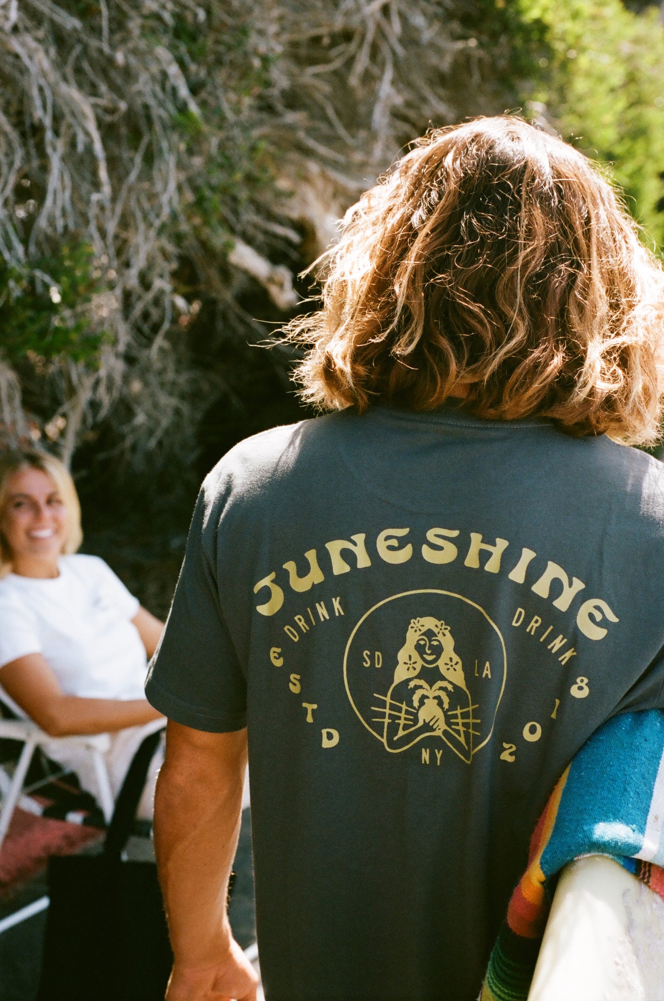 Long haired man wearing a black JuneShine tshirt that reads JuneShine, Estd 2018 in SD, NY, LA with image of a tropical woman holding a pineapple