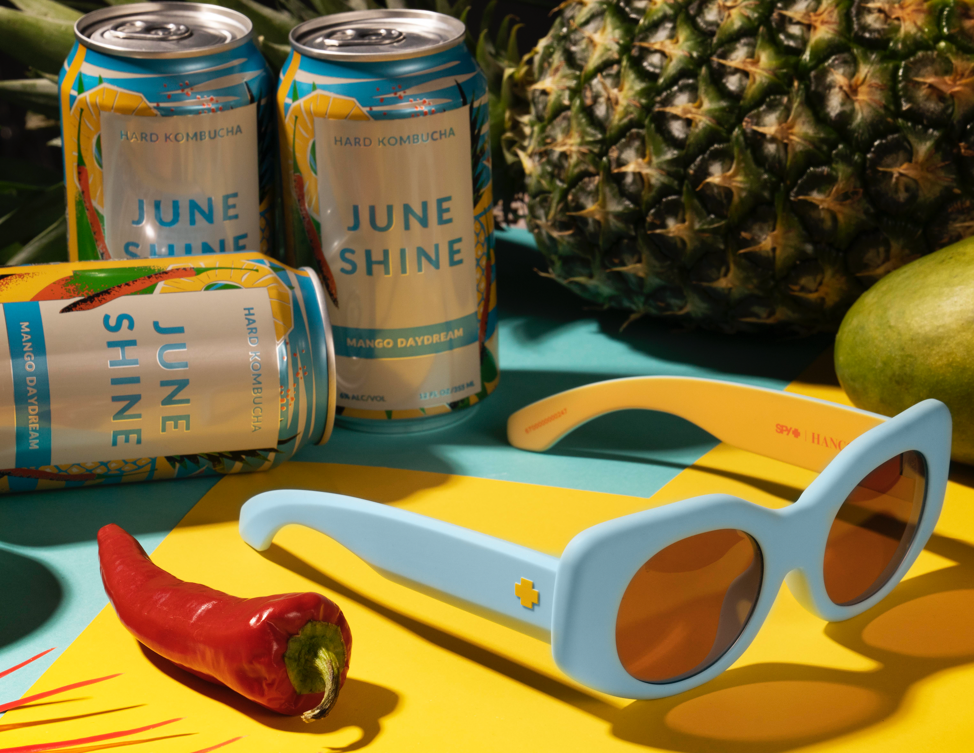 Spy x Juneshine light blue sunglasses against mango daydream cans and pineapple