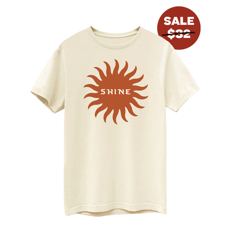 Front view of short sleeve cream colored shirt with a dark orange shape of a sun with the word 