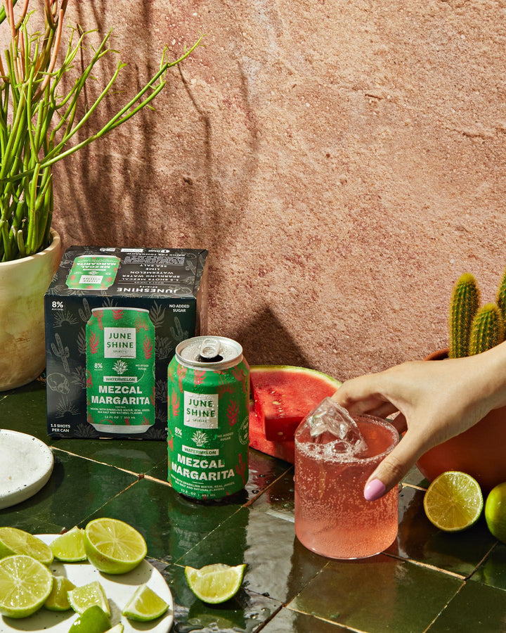 mezcal margarita can and carton with liquid poured into a glass and a hand touching it surrounded by limes and cacti