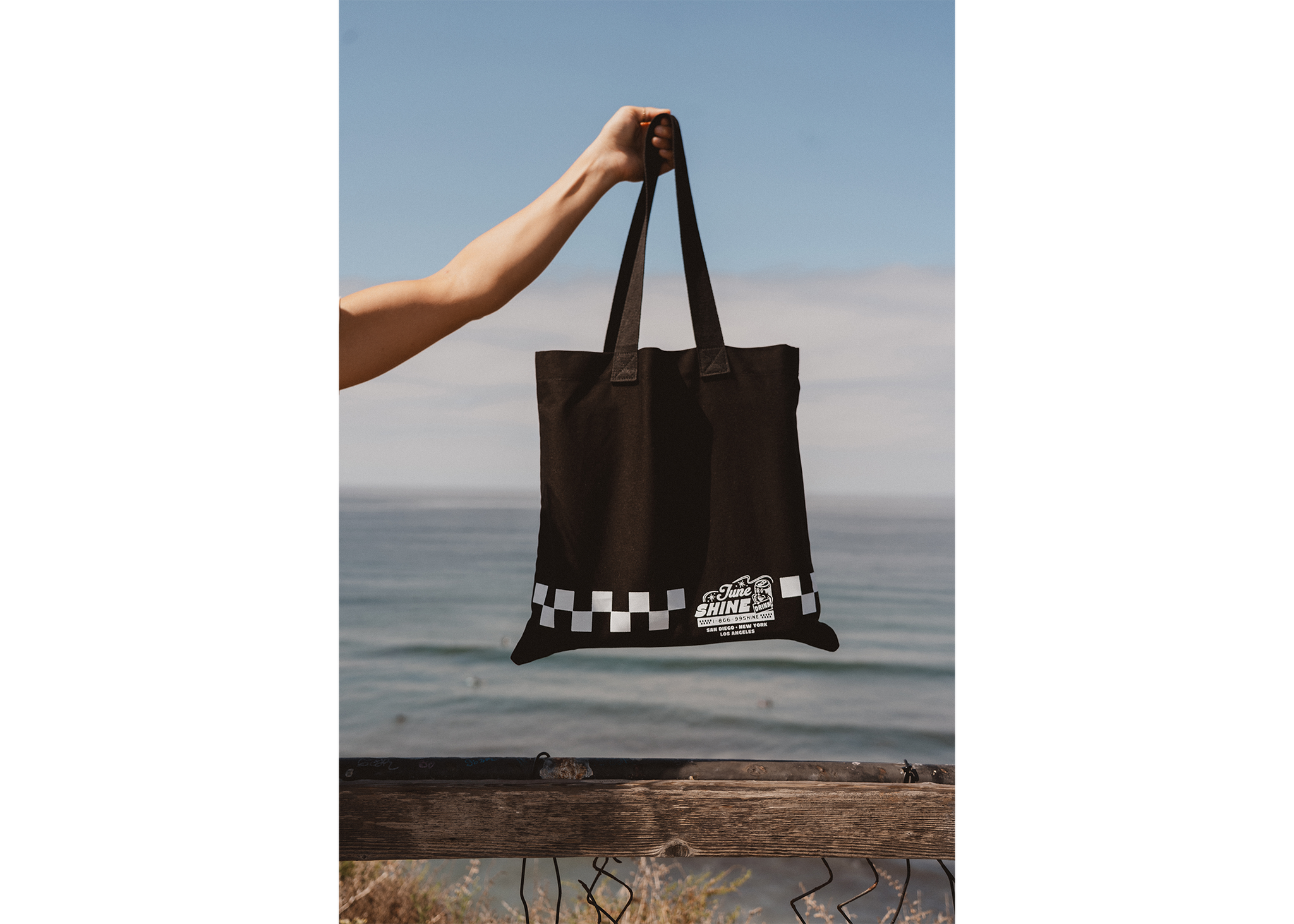 Arm holding Black canvas tote bag with white checkerboard print and text that reads JuneShine, 1-866-99SHINE San Diego New York Los Angeles against an ocean background