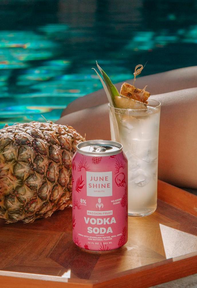 JuneShine Spirits Passion Fruit Vodka Soda can opened staged with garnished cocktail glass, legs and pineapple by a pool.
