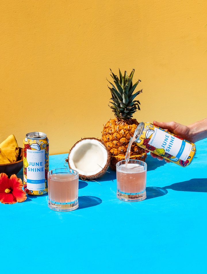 2 coco rico cans, one pouring into a glass with pineapple, coconut, and hibiscus flowers arranged around glasses.