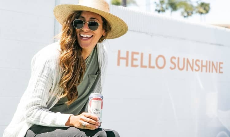 Woman holding JuneShine Acai Berry can wearing a hat a sunglasses with "Hello Sunshine" written on the wall behind her.