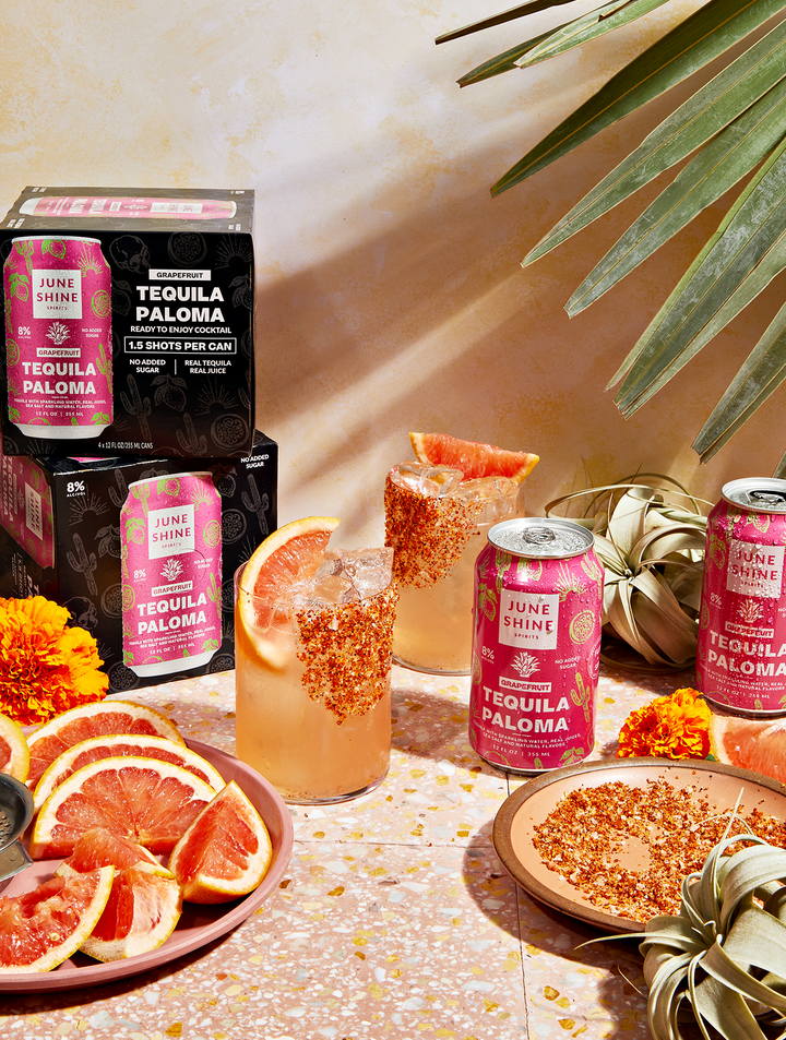 Tequila paloma in cans and poured into glasses over ice with tajin rims and grapefruit slices, surrounded by tajin on plates and grapefruit slides with paloma cartons