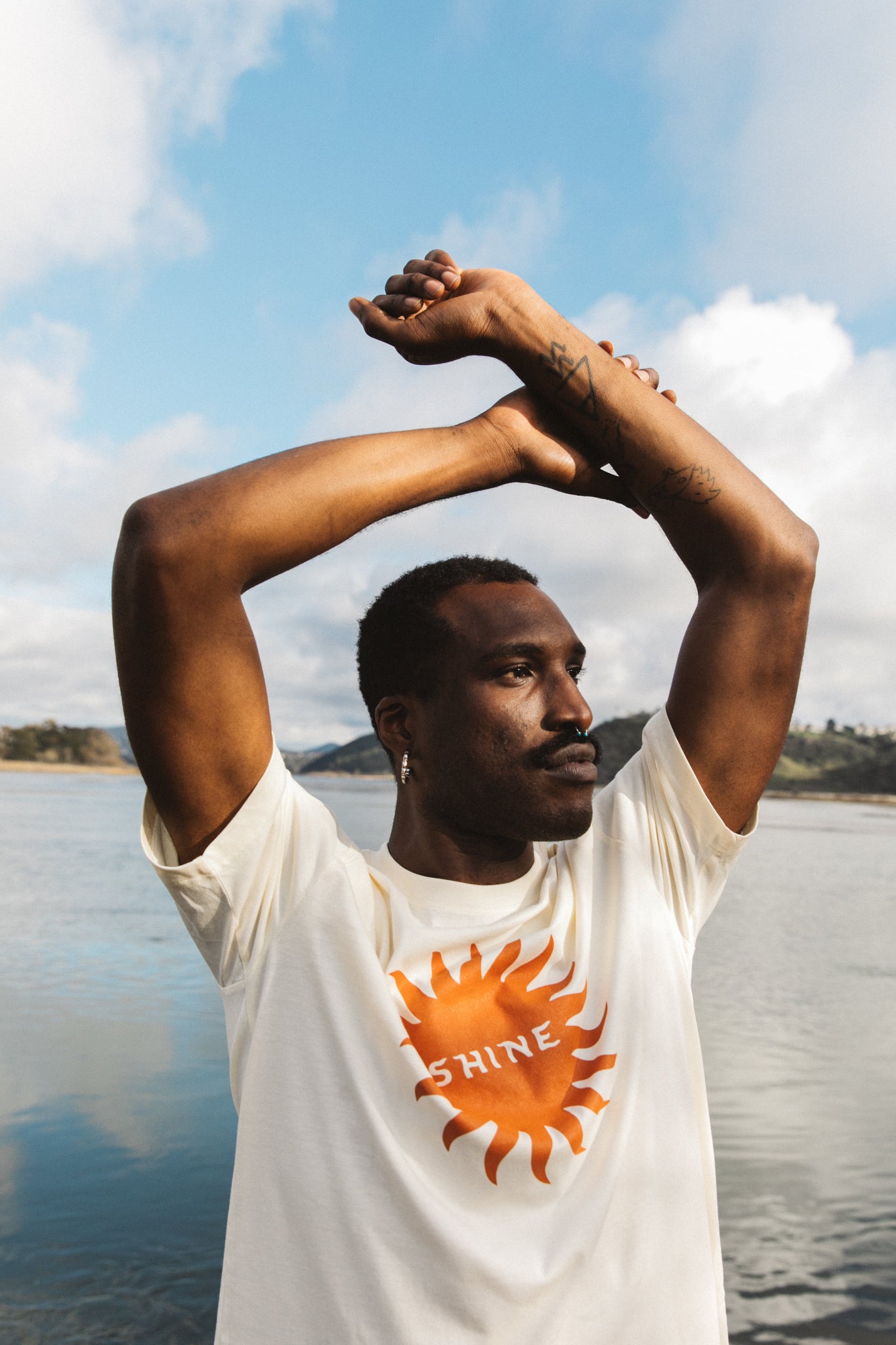 Photograph of man wearing cream colored short sleeve shirt with dark orange sun and word "shine" while having arms in the air.